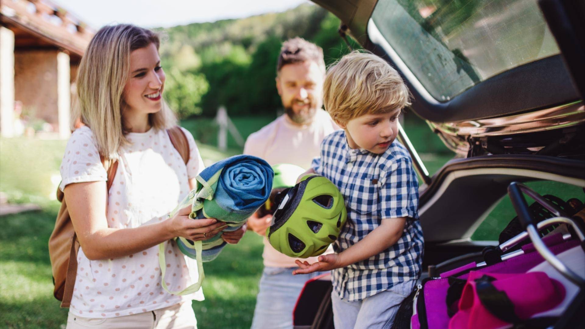 Familie som pakker bilen før en tur, med mor som holder et sammenrullet teppe, far i bakgrunnen og barn som legger en sykkelhjelm i bagasjerommet. Grønne omgivelser og fint vær.